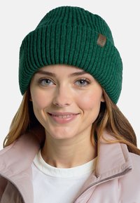Grøn ribbet strikslue med et struktureret mønster og et lille beige lædermærke, båret af en person med lysebrunt hår.