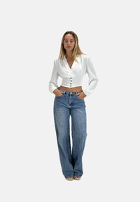 Weißes Croptop mit Wickeldesign und drei Knöpfen vorne. Kombiniert mit hochgeschnittenen, hellblauen, weiten Jeans.