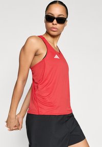 Red athletic tank top with a moisture-wicking fabric, featuring a round neckline and a small white logo. Paired with black shorts.