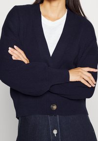 Tmavě modrý žebrovaný cardigan s hlubokým V-výstřihem, dlouhými rukávy a jediným hnědým knoflíkem. Měkká textura a uvolněný střih.