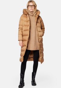 Brązowy puchowy długi płaszcz z wysokim kołnierzem, z poziomymi pikowanymi sekcjami, noszony na beżowym swetrze, czarnych legginsach i botkach za kostkę.