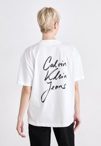 Bijela pamučna majica kratkih rukava, s velikim crnim kurzivnim natpisom "Calvin Klein Jeans" na leđima. Osnovni, ležeran dizajn.