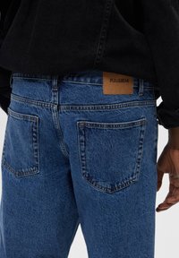 Vaqueros de mezclilla azul con un ajuste recto, que cuentan con dos bolsillos traseros, parche de cuero marrón con la marca y detalles de costura visibles.