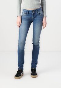 Jeans skinny en denim bleu avec une taille mi-haute, deux poches avant avec des coutures décoratives et une texture lisse. Portés avec des baskets noires.