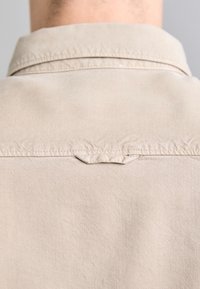 Beige paita, jossa on klassinen kaulus, teksturoitu kangas, ompelukoristeet takana ja selkeä hartialaudekappale.