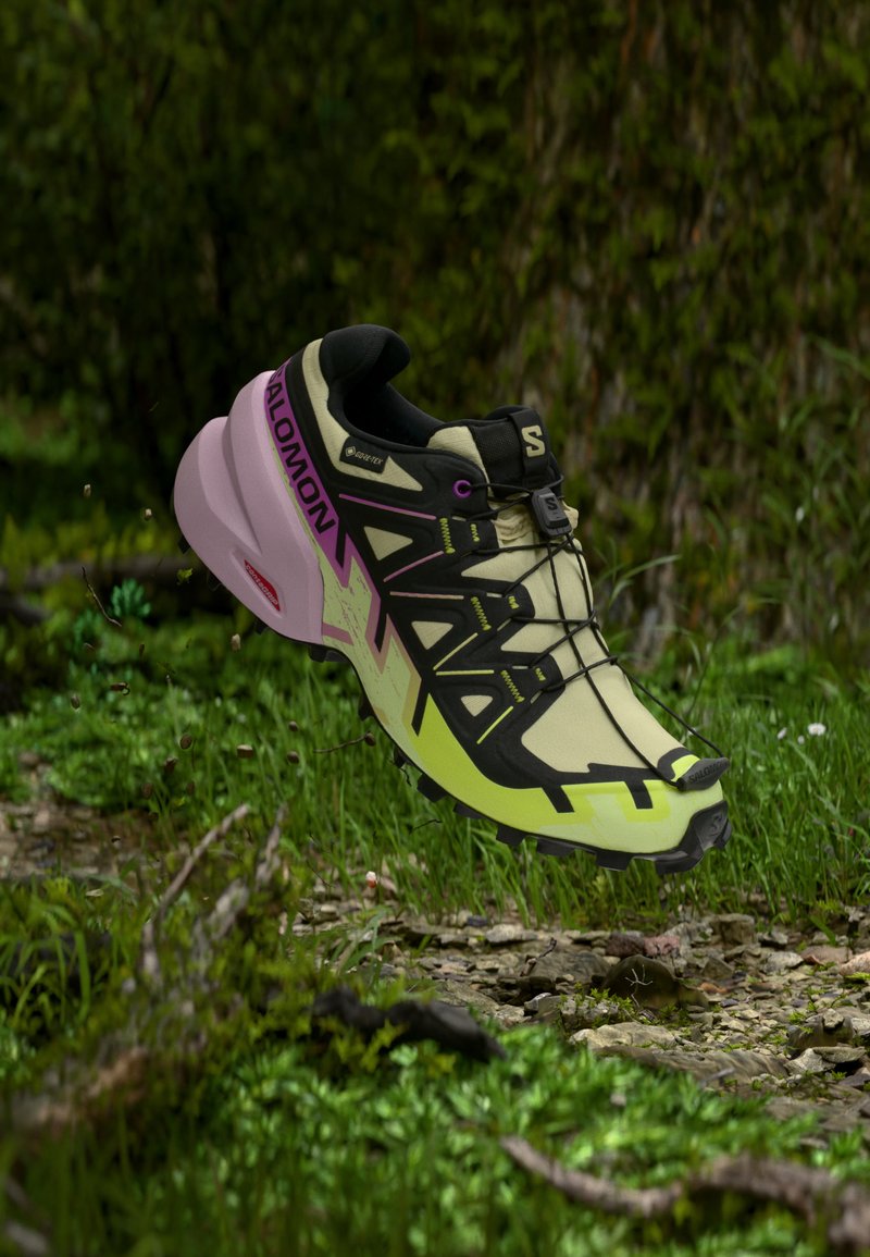 Lys grønn og svart terrengløpesko med lilla såle og hurtiglukkesystem, svever over skogbunn med gress og steiner.