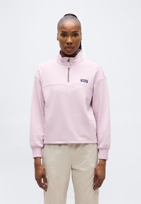 Γυναίκα που φοράει ροζ ανοιχτόχρωμο φλις με φερμουάρ στο στήθος και μπεζ παντελόνι, στέκεται μπροστά σε απλό λ ευκό φόντο.