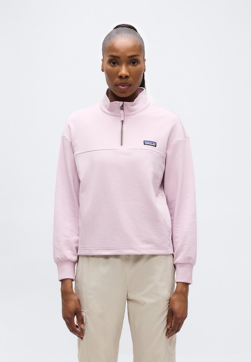 Γυναίκα που φοράει ροζ ανοιχτόχρωμο φλις με φερμουάρ στο στήθος και μπεζ παντελόνι, στέκεται μπροστά σε απλό λευκό φόντο.