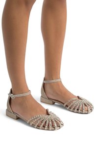 Ballerines beige scintillantes avec un design à lanières, ornées de pierres à rhin. Talon petit et brides de cheville avec une boucle.