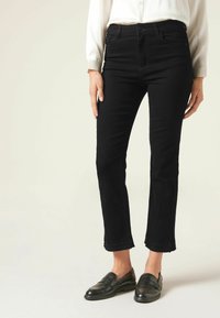 Svarta denimjeans med en tight silhuett, lätt trasighet vid ankelsömmen, i kombination med svarta loafers och en vit skjorta med krage.