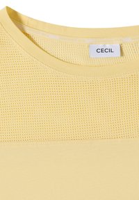 Haut en maille jaune avec un col rond et un détail en mesh sur le haut, avec une étiquette blanche portant le nom de la marque "CECIL" à l'intérieur du col.