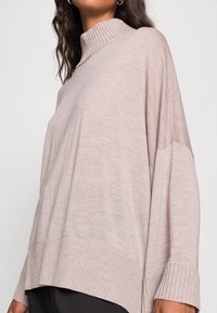 Hellbeiger gestrickter Pullover mit hohem, geripptem Kragen, lockerem Sitz sowie gerippten Bündchen und Saum. Das Material wirkt weich mit einer leicht strukturierten Oberfläche.