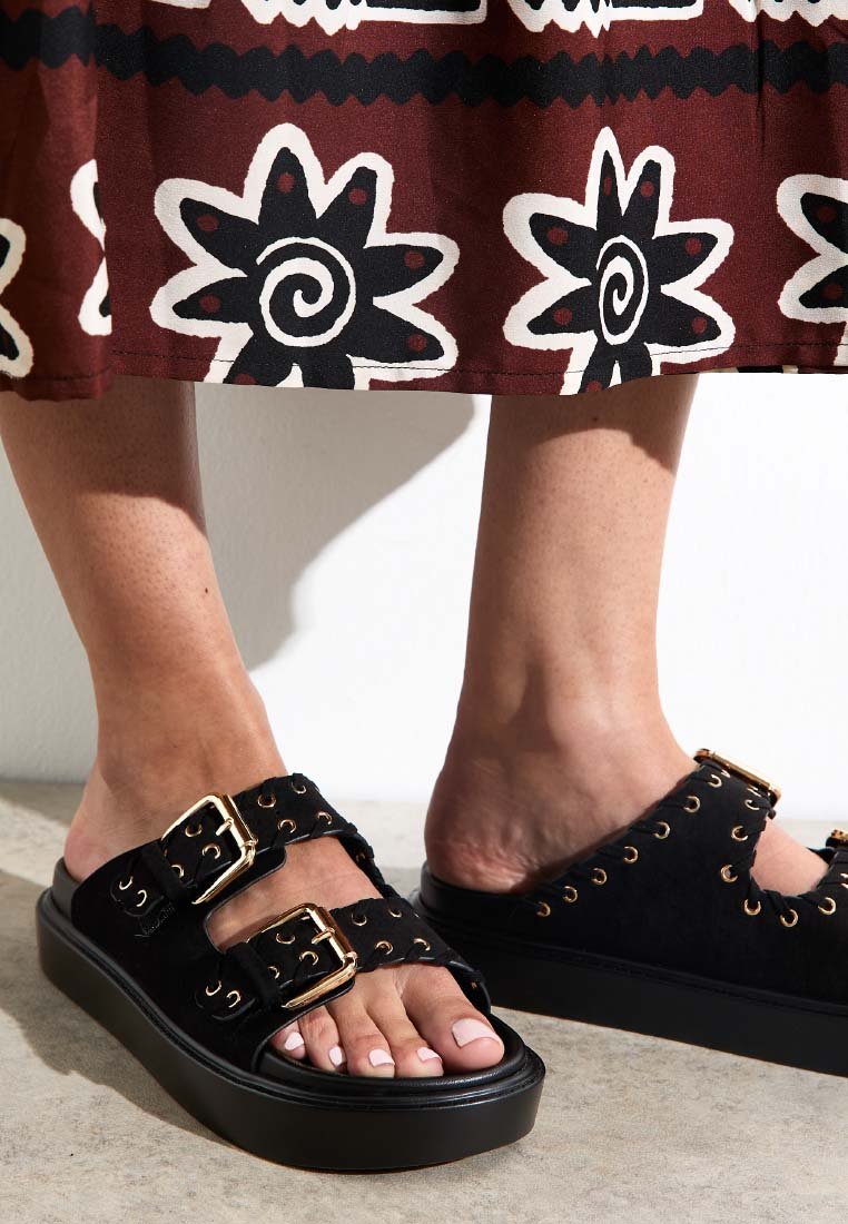 Des sandales en suede noir présentent deux grandes boucles en or, une semelle à plateforme et des découpes. L'arrière-plan montre une jupe à motifs.