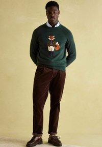 Junger Mann, der einen dunkelgrünen Pullover mit einem Fuchsdesign, braune Cordhosen und dunkelbraune Schuhe trägt, vor einem beigen Hintergrund steht.