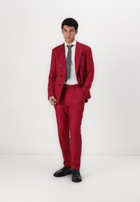 Costum roșu, croit pe măsură, cu sacou cu două rânduri de nasturi și pantaloni slim-fit. Include o cravată gri și o cămașă albă. Pantofii sunt din piele neagră.