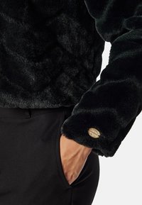 Zwarte nepbont pullover met een gestructureerd oppervlak en een gouden embleem op de mouw. De stof heeft een zachte, luxueuze uitstraling.
