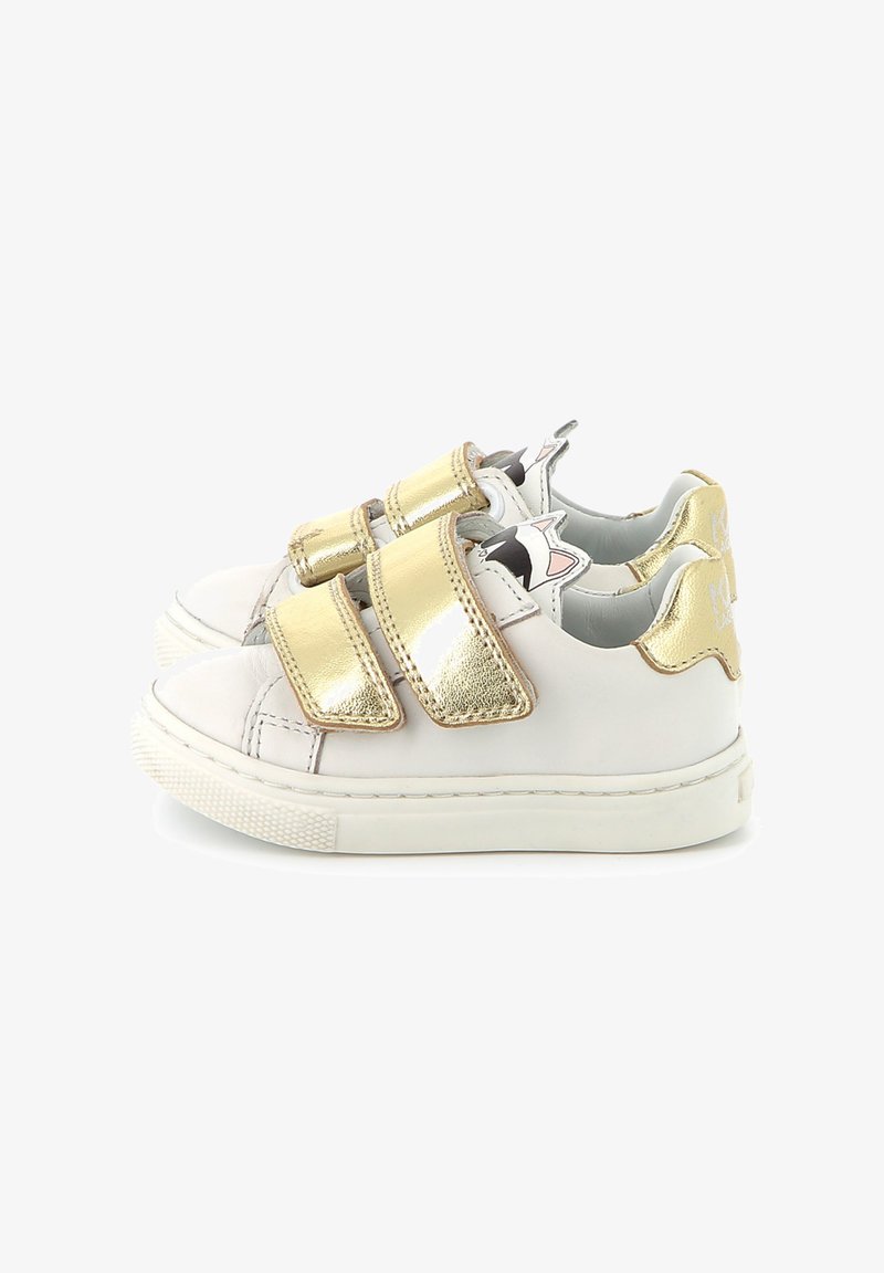 Baskets en cuir blanc avec des sangles velcro métalliques dorées, bout arrondi et semelle en caoutchouc texturé. Présente des accents ludiques sur le talon.