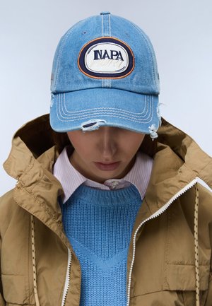 Henkilö, jolla on kulunut sininen farkkulippis, jossa on "NAPA"-logo, sininen neulepusero, raidallinen kauluksellinen paita ja ruskea vetoketjullinen takki.