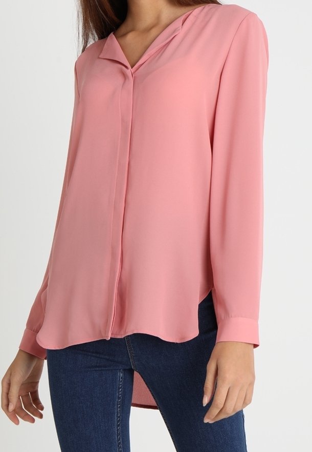 Blouse rose clair à manches longues avec un col en V et une fermeture boutonnée sur le devant. Tissu lisse ; ourlet légèrement arrondi. Portée avec un jean bleu.