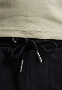 Camiseta verde claro con el dobladillo por encima de pantalones de chándal azul marino oscuros, que presentan un cordón negro con extremos plateados, mostrando materiales texturizados.