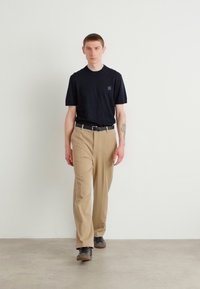 Černé tričko s krátkým rukávem, béžové kalhoty na míru a černý opasek. Model má na sobě černé boty. Jednoduché barvy, minimalistický design, ve stoje.