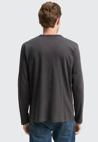 Langärmliges graues Shirt mit rundem Halsausschnitt, von hinten gezeigt, kombiniert mit blauen Jeans, die Details der Stickerei an der Gesäßtasche aufweisen.