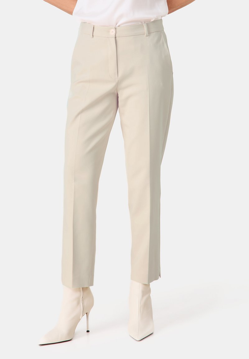 Beige maßgeschneiderte Hosen mit schmaler Passform, weicher Textur und Falten an der Vorderseite, kombiniert mit eleganten weißen spitzen Stiefeletten.
