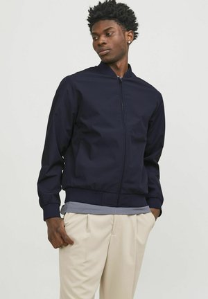 Chaqueta bomber de marina con cierre de cremallera, puños y dobladillo acanalados, llevada sobre una camiseta gris y pantalones de color claro, con una textura suave.