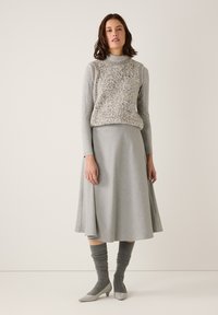 Frau trägt einen grauen Langarm-Rollkragenpullover, eine Weste mit Leopardenmuster, einen Midirock, graue lockere Socken und hellgraue niedrige Schuhe, steht vor einer beigen Wand.