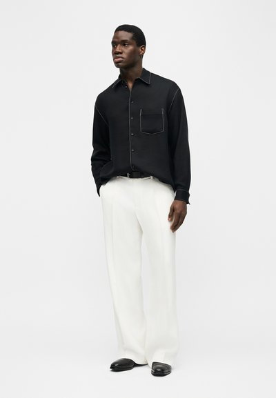 Άνδρας που φοράει μαύρο πουκάμισο με κουμπιά και λευκή ραφή, λευκό παντελόνι με φαρδιά πόδια, μαύρη ζώνη και μαύρα παπούτσια, στέκεται μπροστά σε λευκό φόντο.