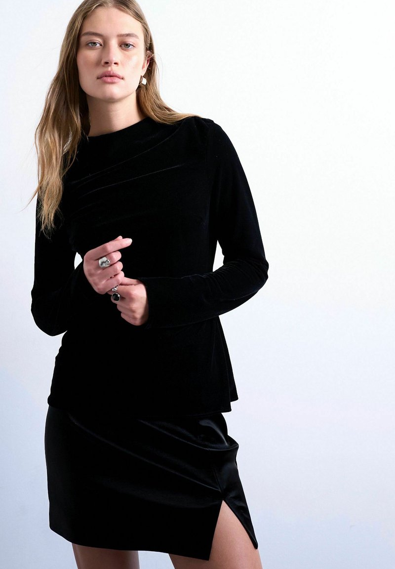 Femme aux longs cheveux blonds portant un haut à manches longues en velours noir et une jupe noire fendue sur le côté, sur fond blanc.