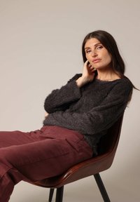 Fluffy zwart trui met lange mouwen, gecombineerd met bordeauxrode broek, zittend op een bruine stoel met schuine poten tegen een eenvoudige achtergrond.