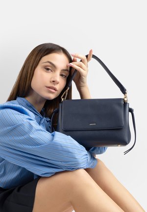 Giovane donna con camicetta a righe blu che tiene una borsa a tracolla in pelle blu navy con dettagli dorati su sfondo bianco.