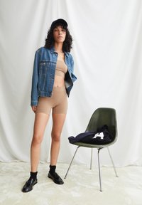 Veste en denim, crop top beige et short de cycliste. Baskets noires et chaussettes. Chaise verte avec un sweat à capuche bleu marine posé dessus. Fond blanc.
