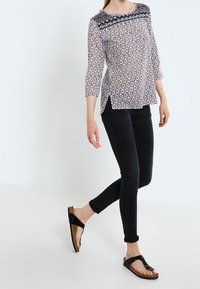 Blouse légère à motifs avec encolure ronde, manches trois-quarts, accent marine aux épaules, associée à un jean noir slim et des sandales noires.