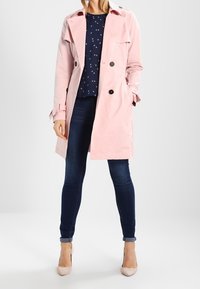 Jasnoróżowy płaszcz typu trencz o luźnym kroju, z podwójnym rzędem guzików i bocznymi kieszeniami, zestawiony z ciemnoniebieskimi dżinsami i beżowymi szpilkami.