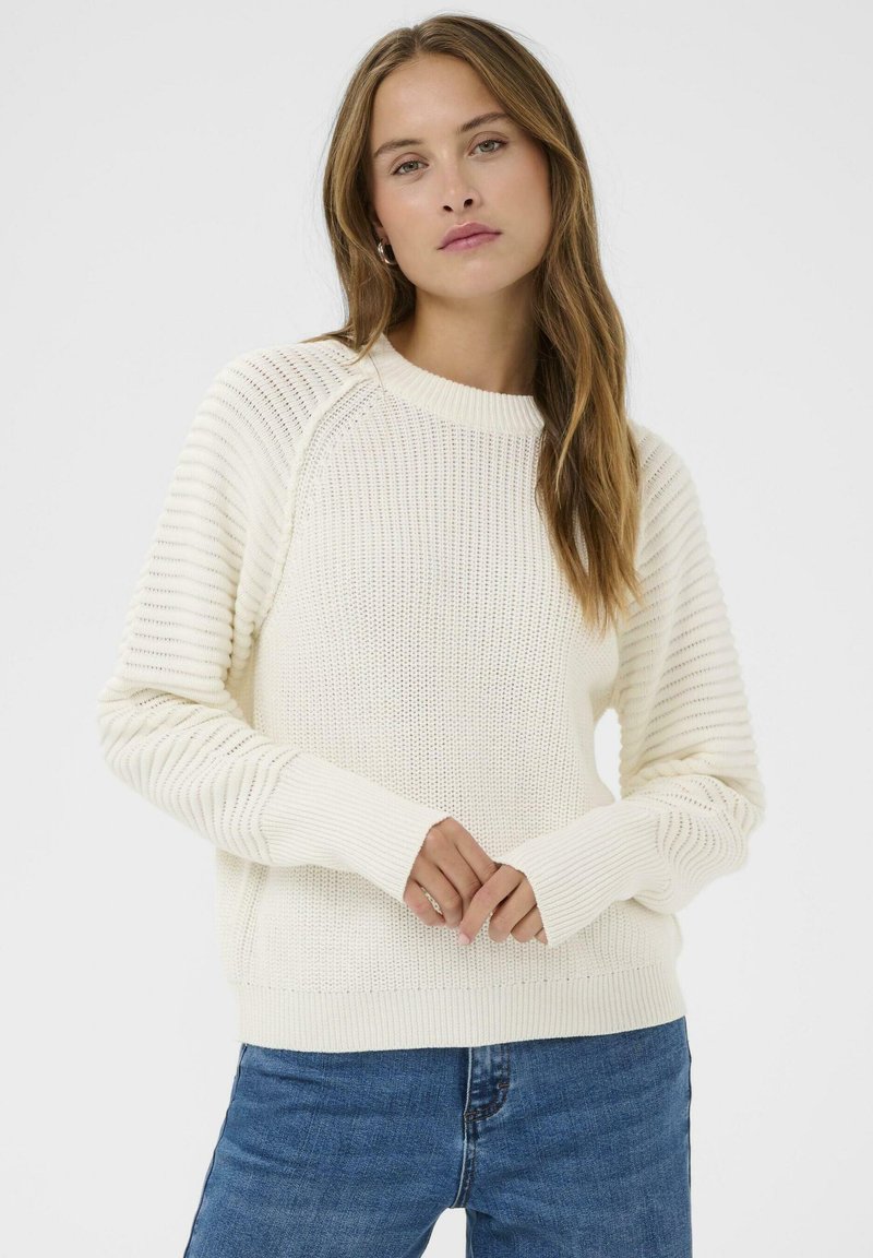 Femme dans un pull en tricot blanc avec des manches texturées et un jean bleu, se tenant devant un fond clair et uni.