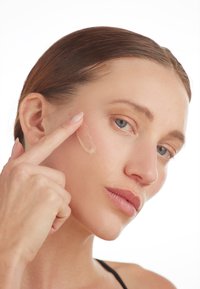 Nærbilde av en person som påfører en kremet foundation på kinnet. Huden har en jevn tekstur, med en lys beige farge som blander seg sømløst.