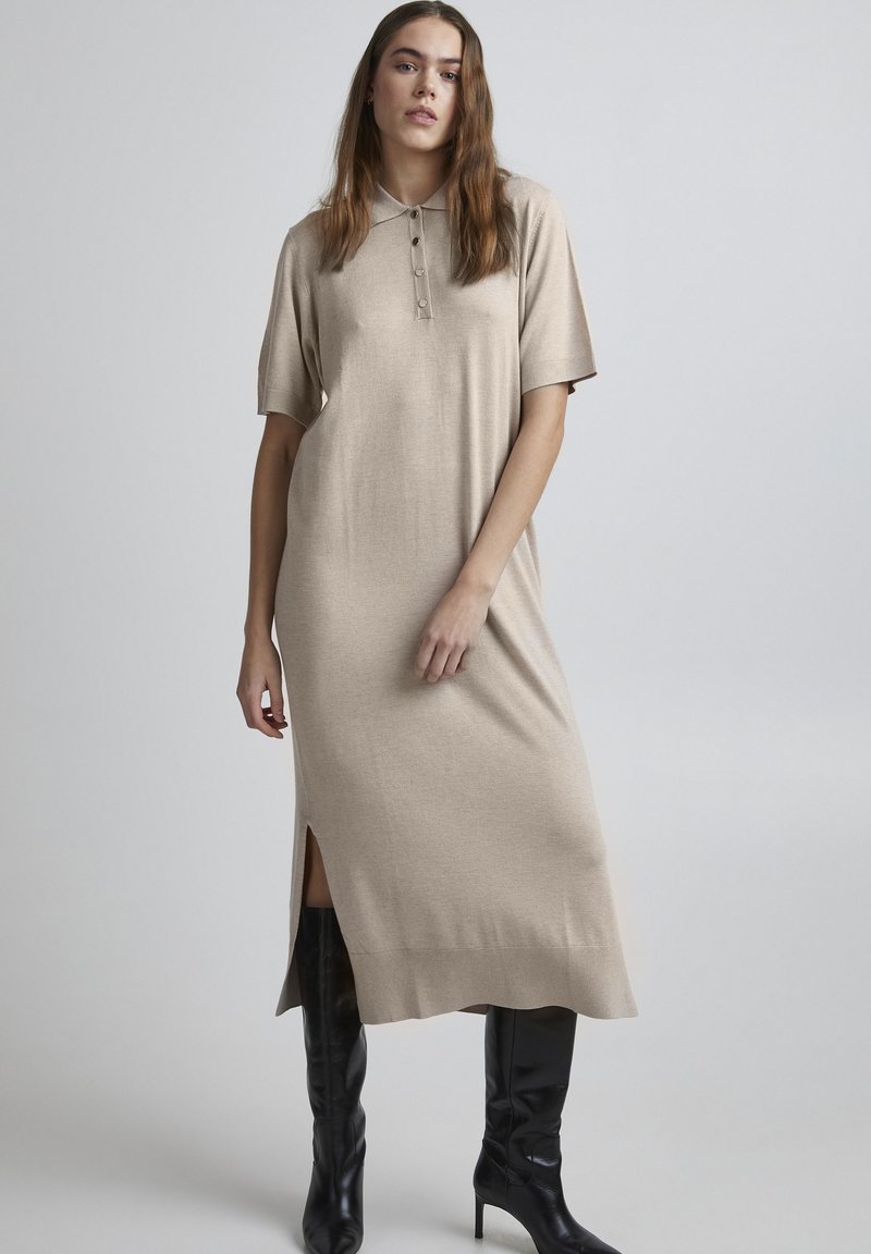Beige strikket kjole med korte ærmer, en krave og knapdetaljer. Har en sideslids og en afslappet pasform, parret med sorte knæhøje støvler.