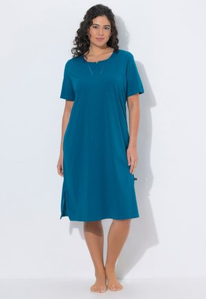 Mujer está descalza apoyada contra una pared blanca, lleva un vestido azul verdoso hasta la rodilla, de manga corta, con pequeñas aberturas laterales y un escote en forma de muesca.