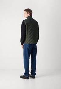 Gilet verde trapuntato con colletto alto, indossato sopra una maglietta nera a maniche lunghe, abbinato a jeans blu e scarpe nere, visto da dietro.