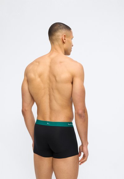 Μυώδης άντρας από πίσω, φοράει μαύρα αντρικά εσώρουχα boxer με πράσινη μέση, μπροστά σε απλό λευκό φόντο.