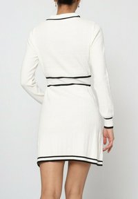 Robe blanche à manches longues, en tissu texturé, avec des rayures noires au niveau du col, de la taille et des poignets. Longueur mi-cuisse.