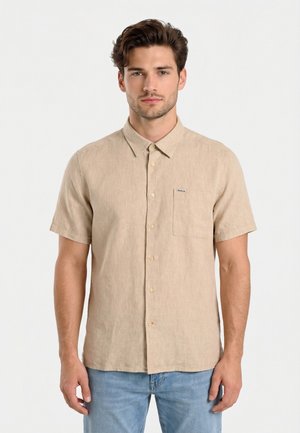 Jonge man met kort bruin haar, gekleed in een beige overhemd met korte mouwen en lichtblauwe spijkerbroek, staand tegen een witte achtergrond.