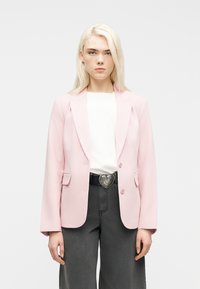 Νεαρό άτομο με μακριά ξανθά μαλλιά που φοράει ανοιχτό ροζ σακάκι, λευκό πουκάμισο, σκούρα γκρι παντελόνια και μαύρη ζώνη με αγκράφα σε σχήμα καρδιάς.