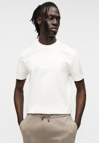 Λευκό βαμβακερό μπλουζάκι με κοντά μανίκια και ανάγλυφο λογότυπο Aston Martin. Συνδυασμένο με μπεζ παντελόνι με κορδόνι. Ομαλή υφή.