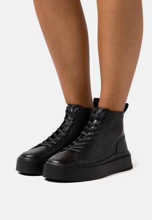 Czarne skórzane sneakersy na wysokiej platformie z sznurowadłami, noszone na gołych nogach na jednolitym jasnym tle.