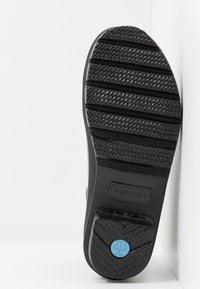 Semelle en caoutchouc noir avec un motif d'adhérence texturé. Présente le logo "HUNTER" en relief et un accent circulaire bleu près du talon.