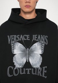 Melnā kapuce ar lielu pelēku tauriņu grafiku un "VERSACE JEANS COUTURE" tekstu sudrabkrāsā. Izgatavota no mīkstas auduma, brīva piegriezuma.