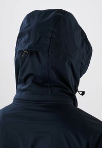 Personne portant une veste noire à capuche avec des cordons réglables à l'arrière de la capuche, tournée dos à la caméra.